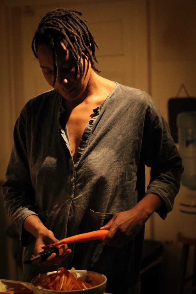 V Woods stands in a warm kitchen, wearing a grey tunic, peeling carrots into a silver bowl